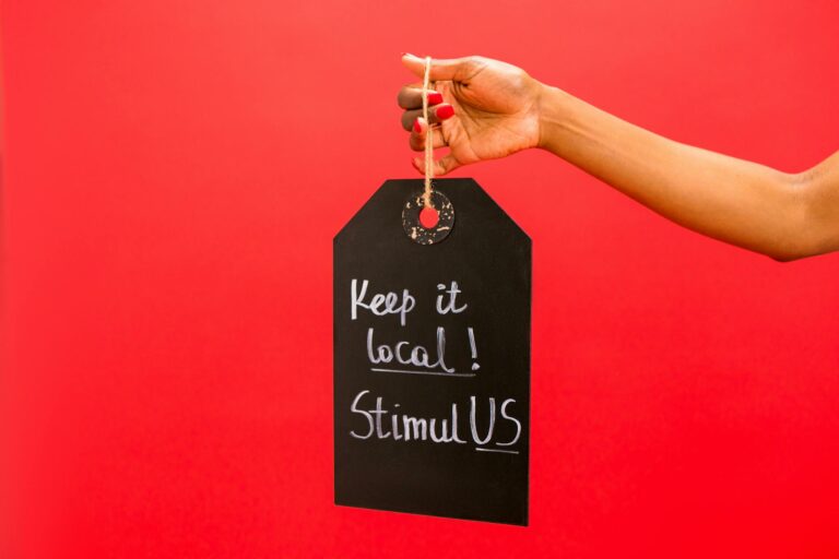 A hand presenting a chalkboard tag reading 'Keep it local! StimulUS' against a vibrant red backdrop.